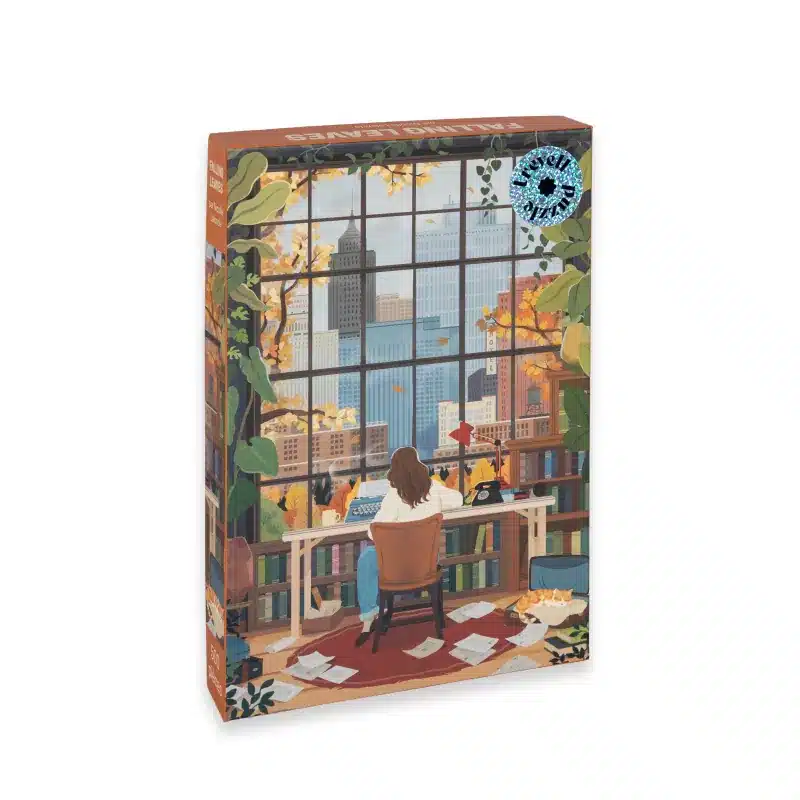Puzzle Falling Leaves Trevell – 500 pièces, illustration d’une jeune fille assise à son bureau écrivant près d’une fenêtre avec vue sur la ville aux couleurs d’automne.